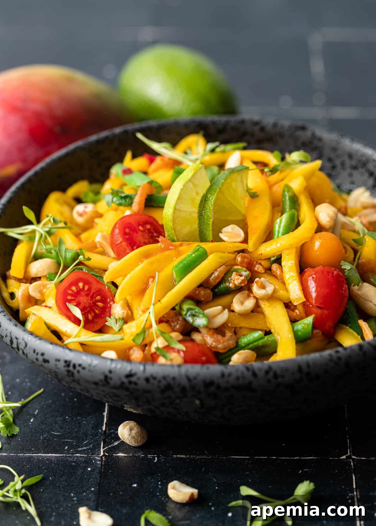 Side view of a serving bowl filled with vibrant green mango salad, featuring crisp green beans, roasted peanuts, and halved cherry tomatoes.