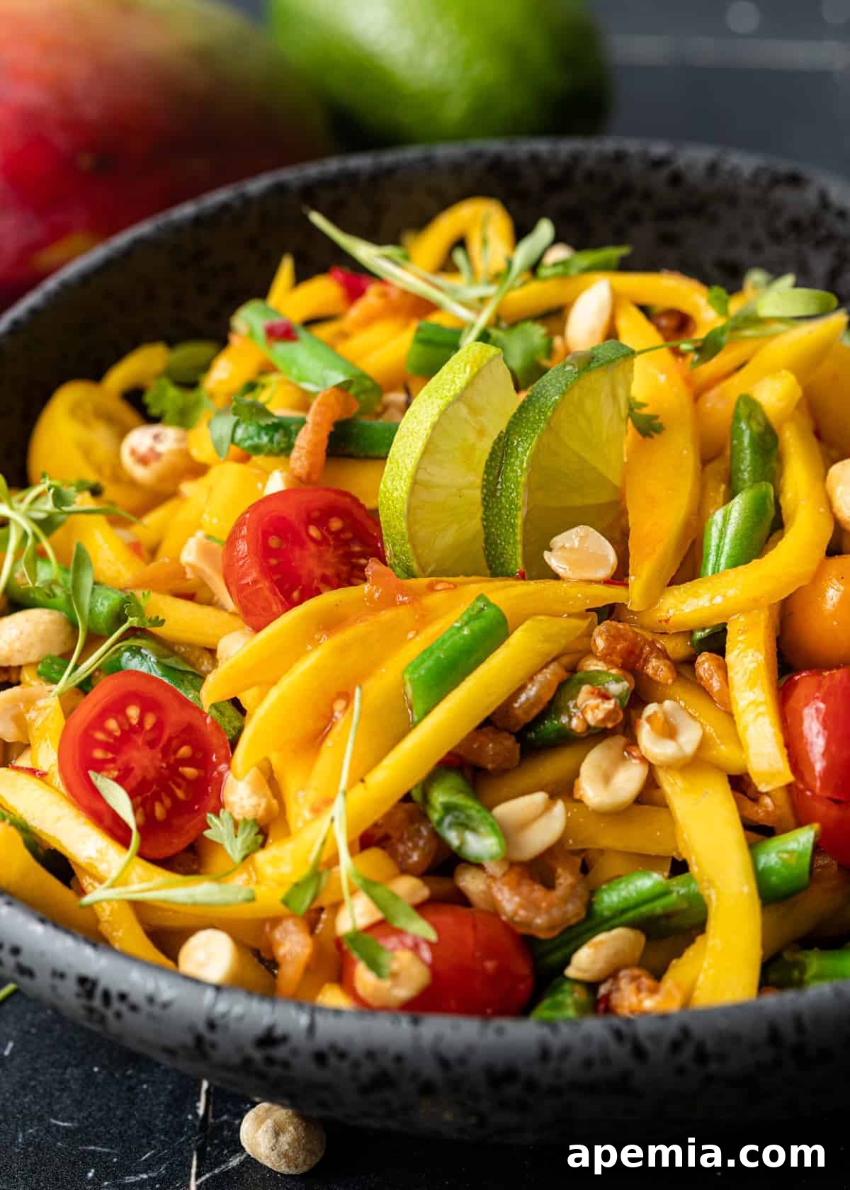 Close-up side view of a vibrant green mango salad in a black bowl, showcasing the julienned mango, crisp green beans, cherry tomatoes, and scattered peanuts.