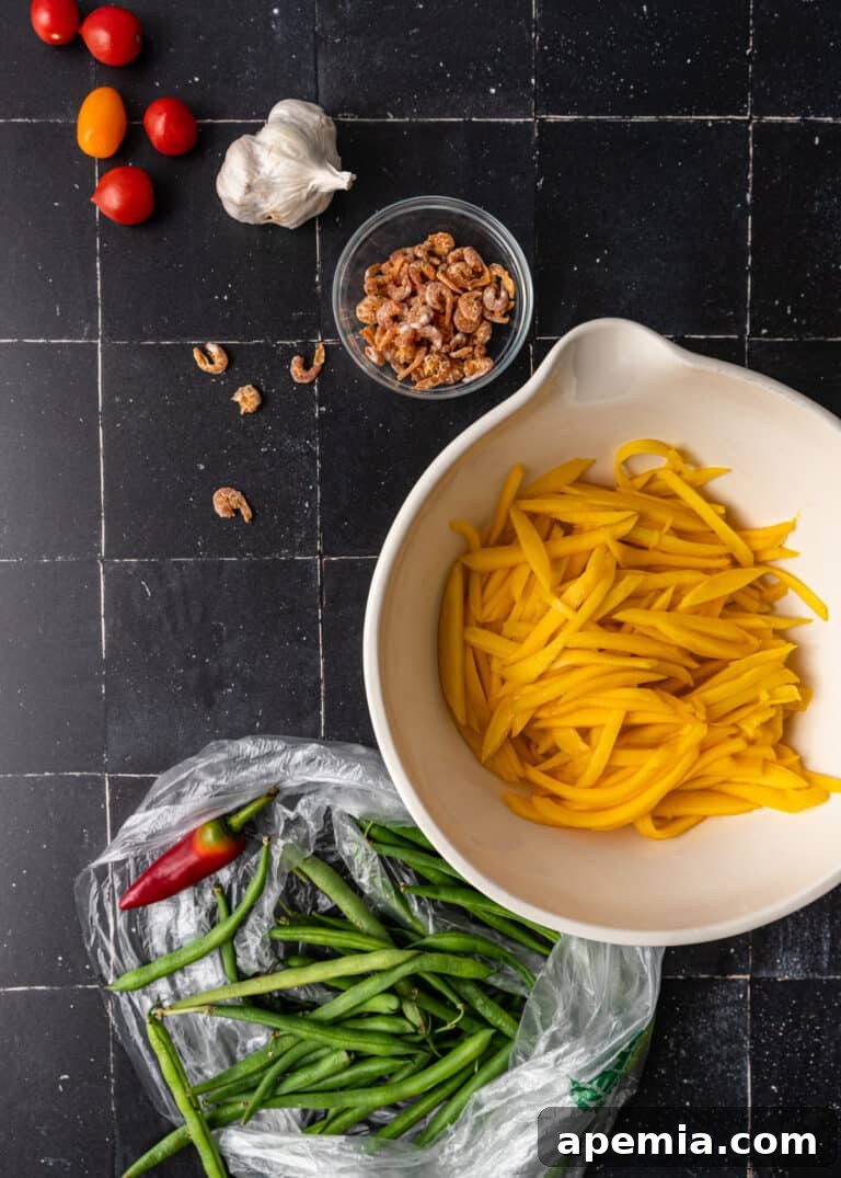 Assorted ingredients for an Asian salad recipe arranged on a black countertop: sweet mango fruit, fresh green beans, dried shrimp, garlic, and cherry tomatoes.