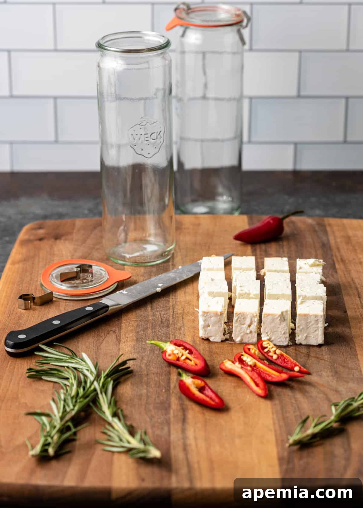 large wooden cutting board with cubes of fresh feta, rosemary sprigs, red chili peppers, and ingredients for marinating cheese