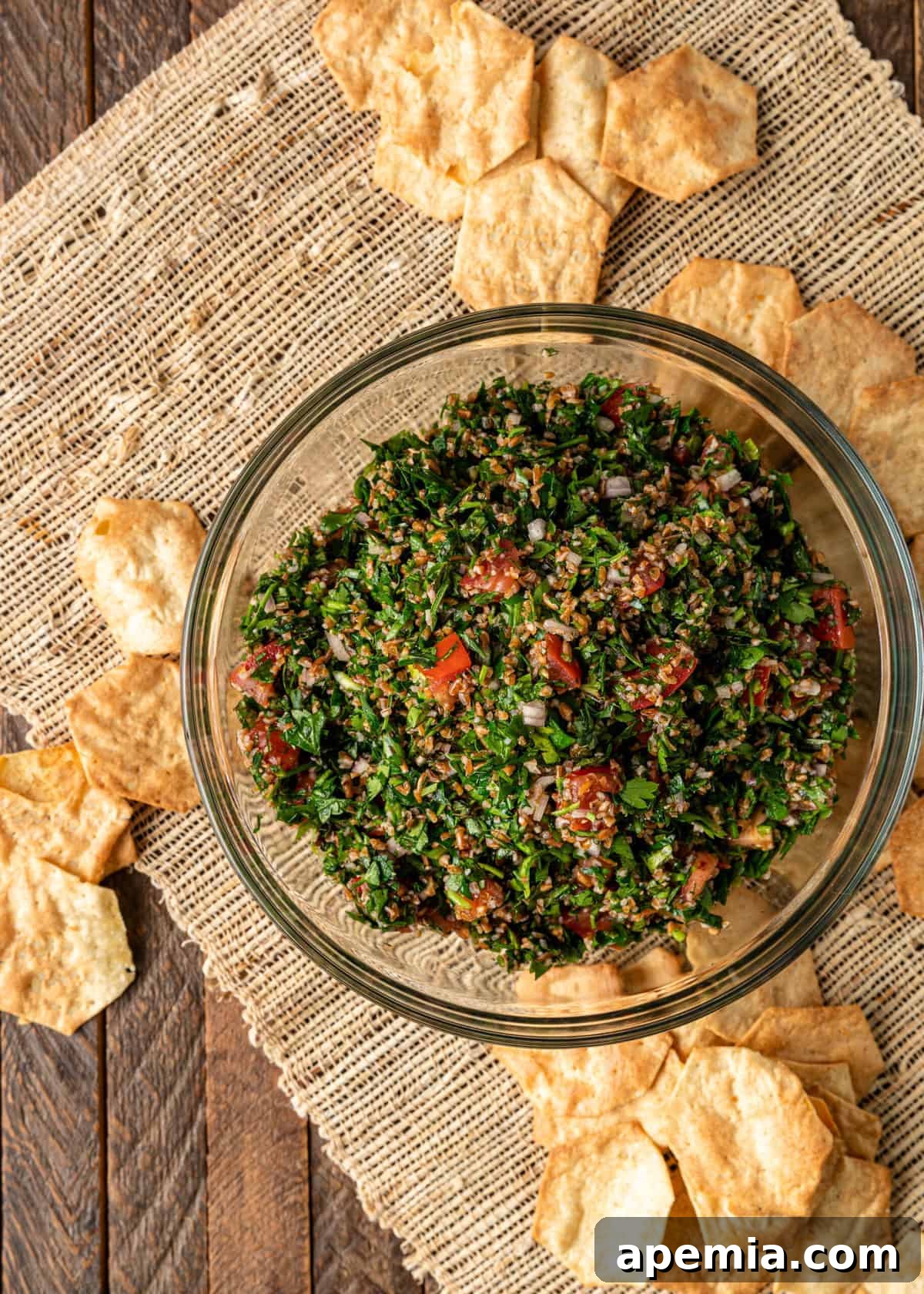 Overhead view of healthy bulgur and parsley tabouli salad in a clear glass bowl, surrounded by crispy pita chips.