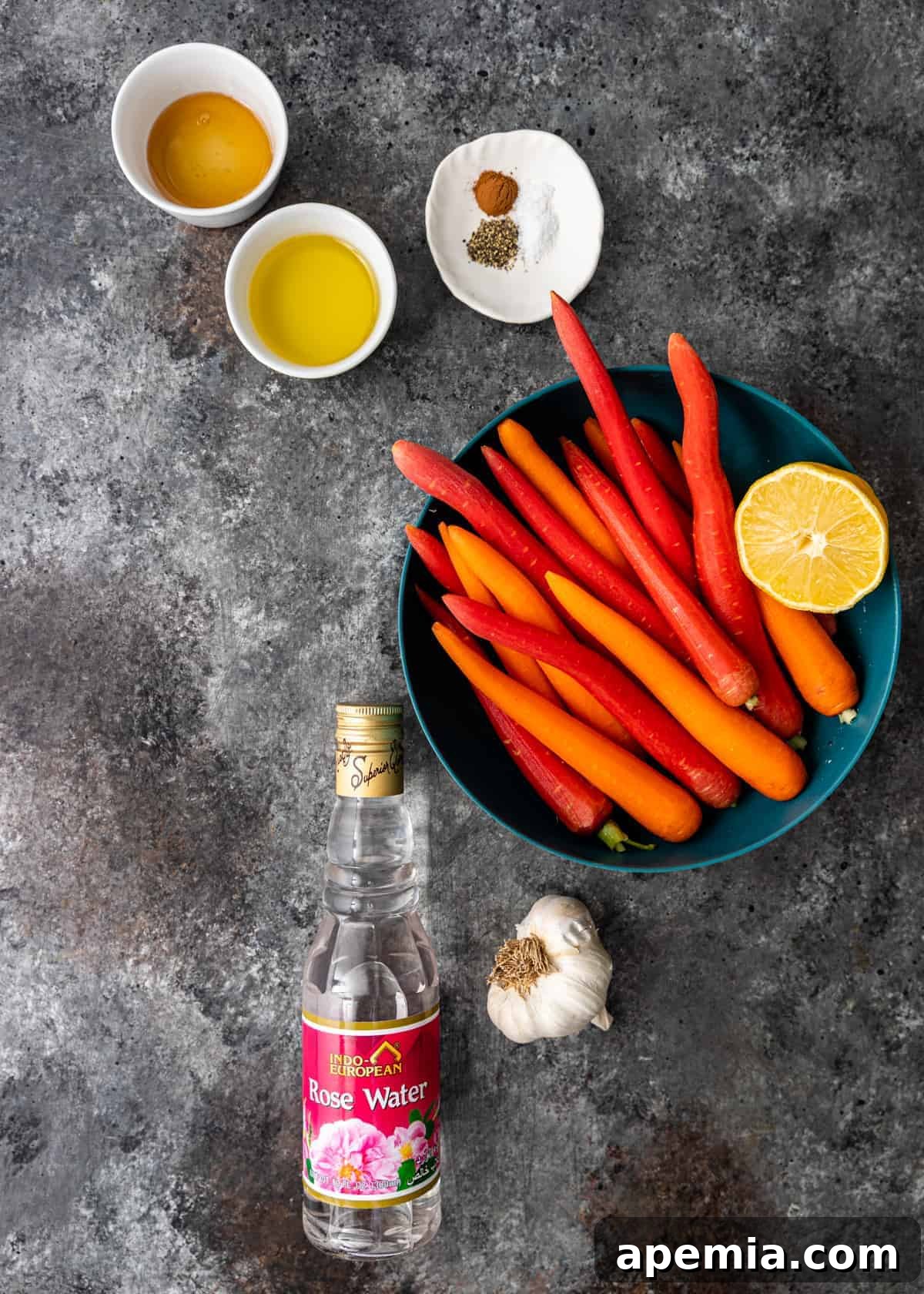 Overhead shot of all ingredients for the baby carrot recipe: fresh heirloom baby carrots, a bottle of rose water, fresh garlic, a lemon, and a selection of spices including cinnamon.