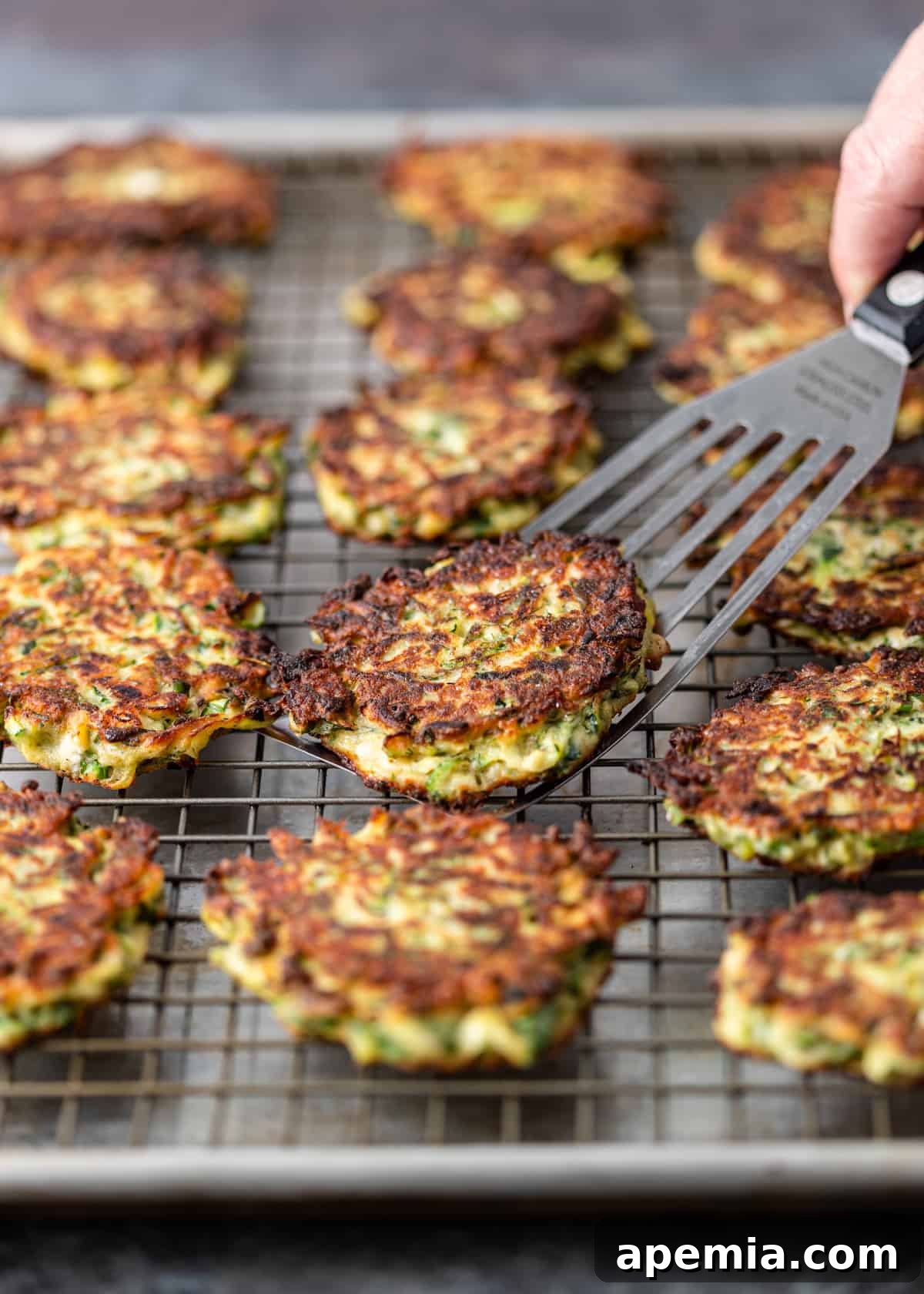 sliding metal spatula under golden brown crispy pan fried zucchini fritters on metal baking rack