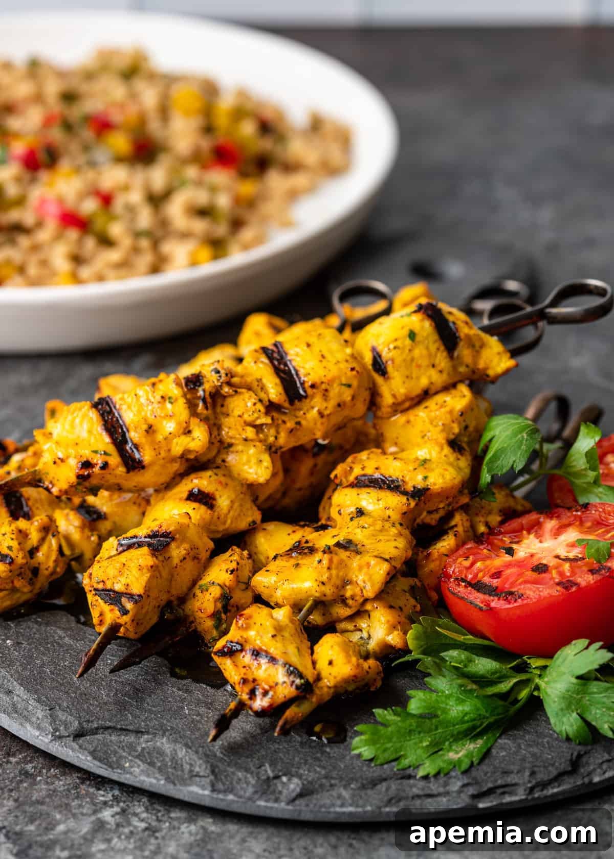 A beautifully arranged plate featuring my Joojeh Kabob recipe with expertly grilled tomatoes and fresh herbs. A bowl of fluffy couscous salad with sautéed vegetables sits invitingly in the background, completing a delightful meal.