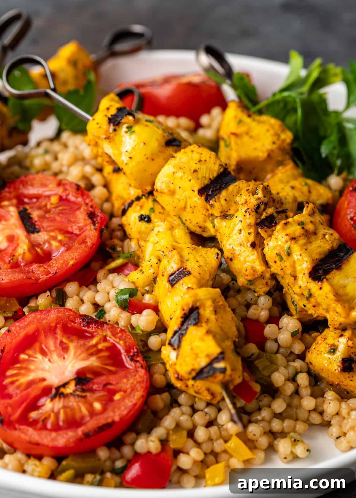 Expertly grilled Persian Joojeh Kabob served over a bed of fluffy couscous, adorned with vibrant grilled tomatoes and fresh herbs, ready to be enjoyed.