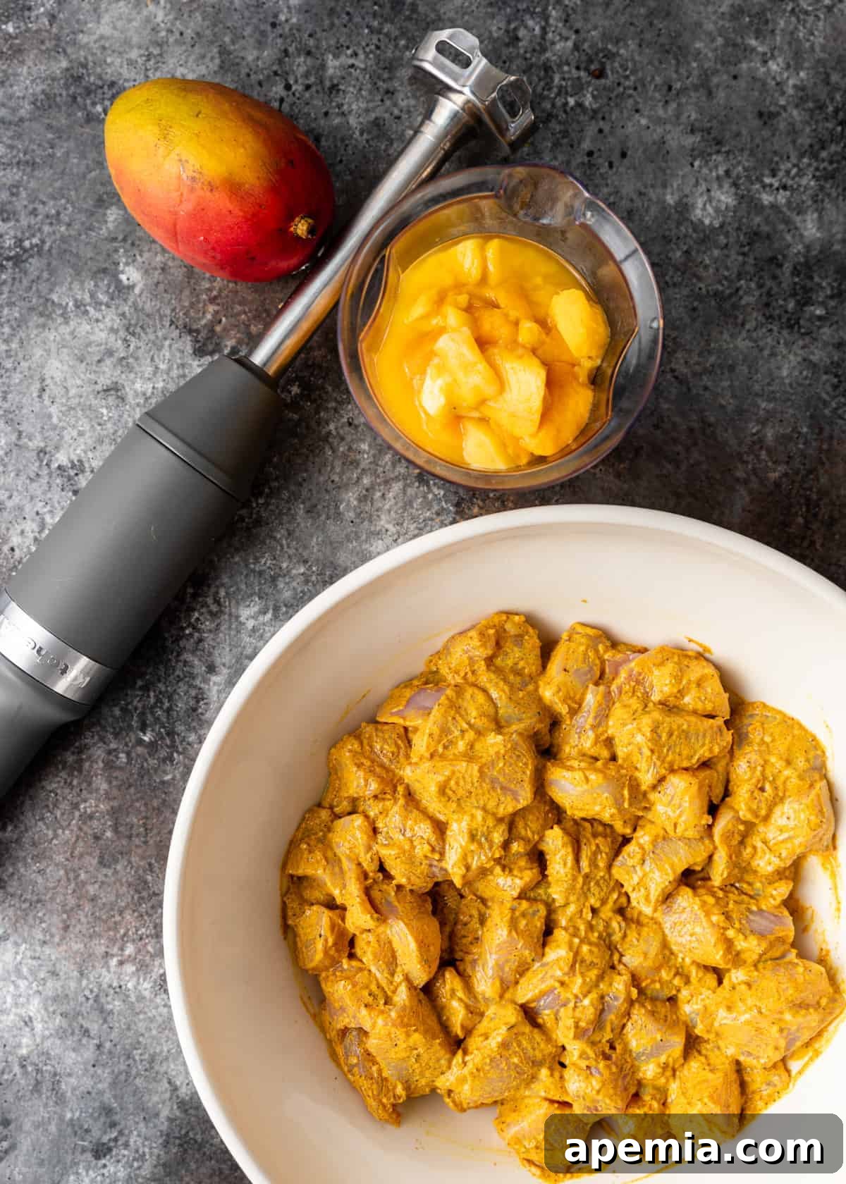 Overhead shot of diced chicken breast in a bowl with yellow marinade, next to a bowl of mango puree and an immersion blender