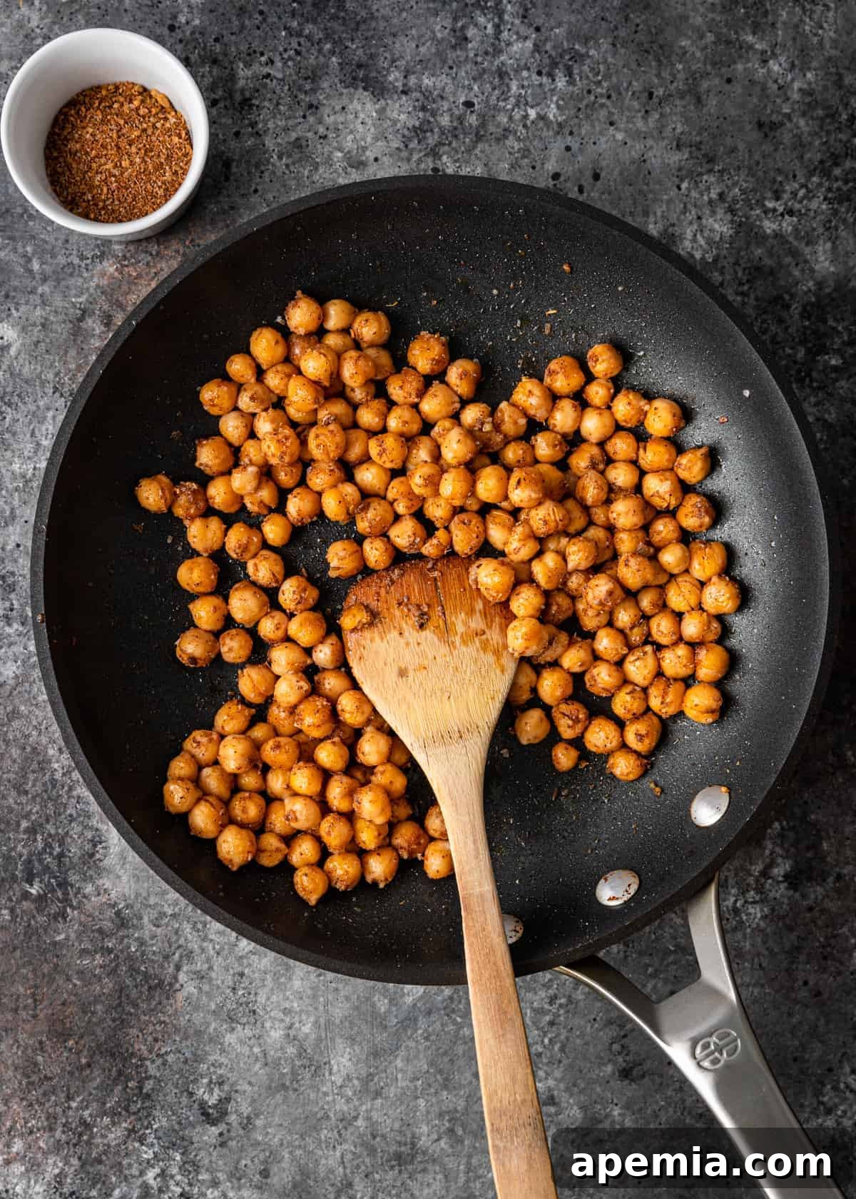 A close-up overhead view of garbanzo beans (chickpeas) being toasted in a skillet, seasoned with spices, to achieve a crispy texture for Mediterranean pizza.