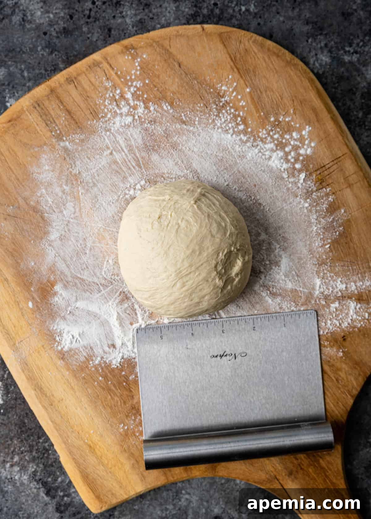 Overhead view of a perfectly shaped ball of homemade pizza dough resting on a floured wooden board, ready for stretching and pan-frying.