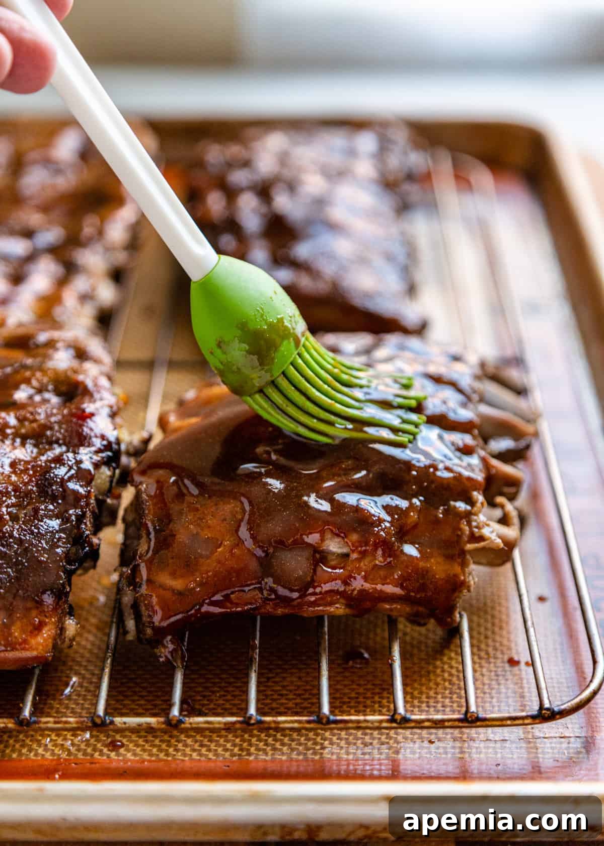 Green silicone basting brush applying glistening sweet and sour sauce to ribs on a baking sheet, ready for broiling.