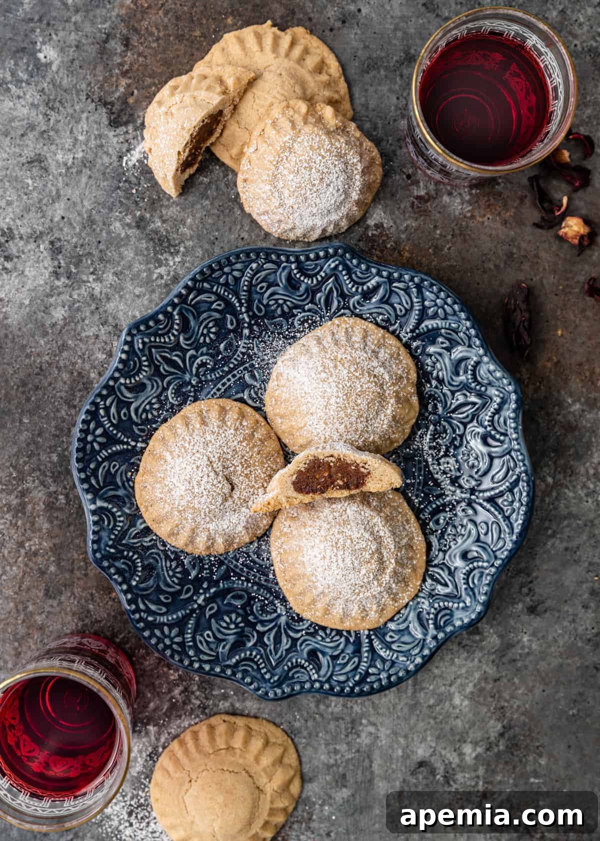 Overhead shot of freshly baked date-filled maamoul cookies arranged on a small blue plate, with more cookies stacked in the background on a kitchen counter.