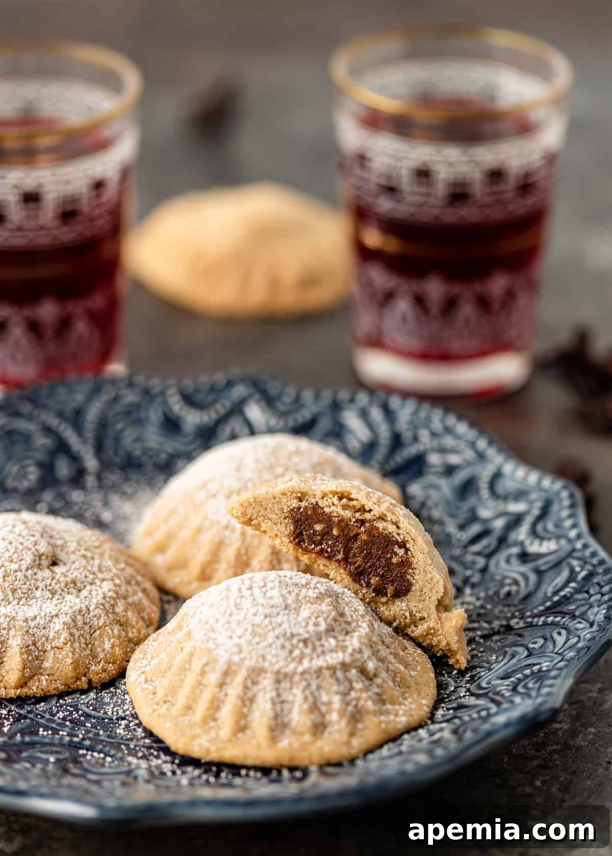 Four golden-brown maamoul cookies delicately placed on a blue plate, accompanied by glasses of vibrant hibiscus tea in the background, suggesting a delightful serving suggestion.