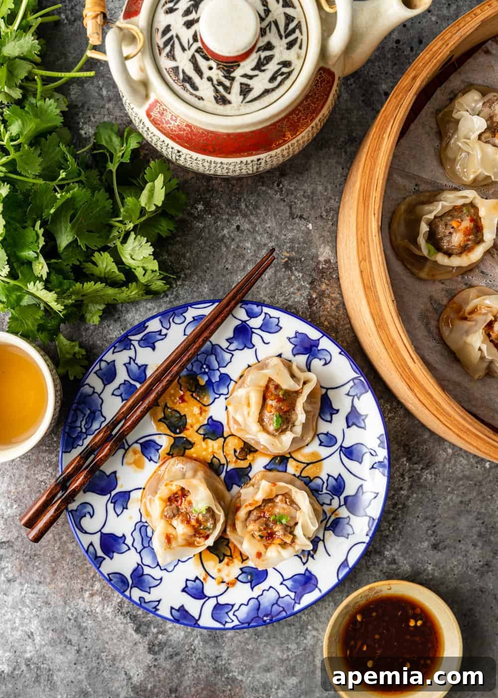 overhead photo of pork shumai on a plate and in a steamer basket with a pot of tea and a bunch of cilantro