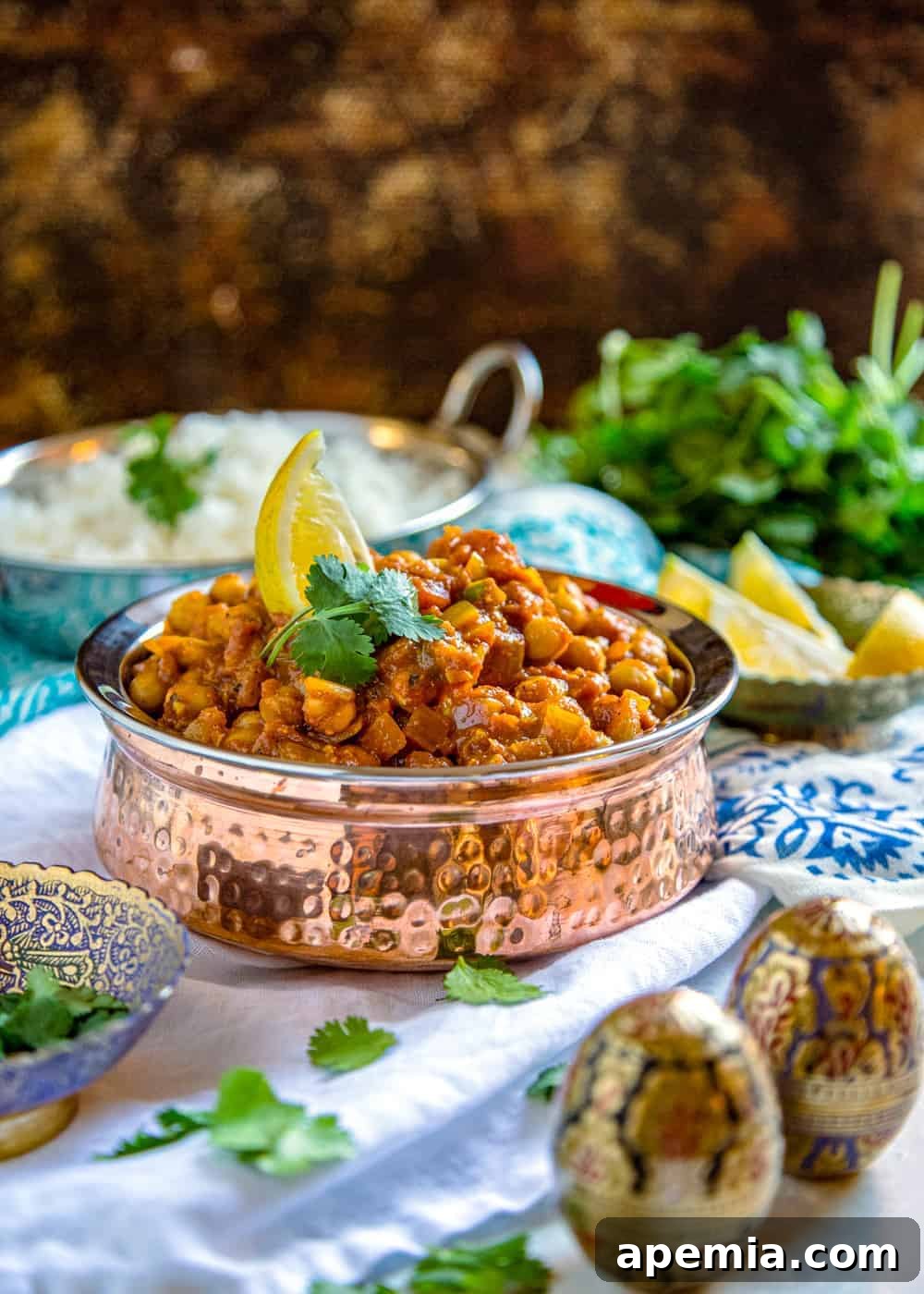 Side view of a rustic copper serving dish, full of homemade chickpea masala, adorned with vibrant fresh cilantro and a slice of lemon.