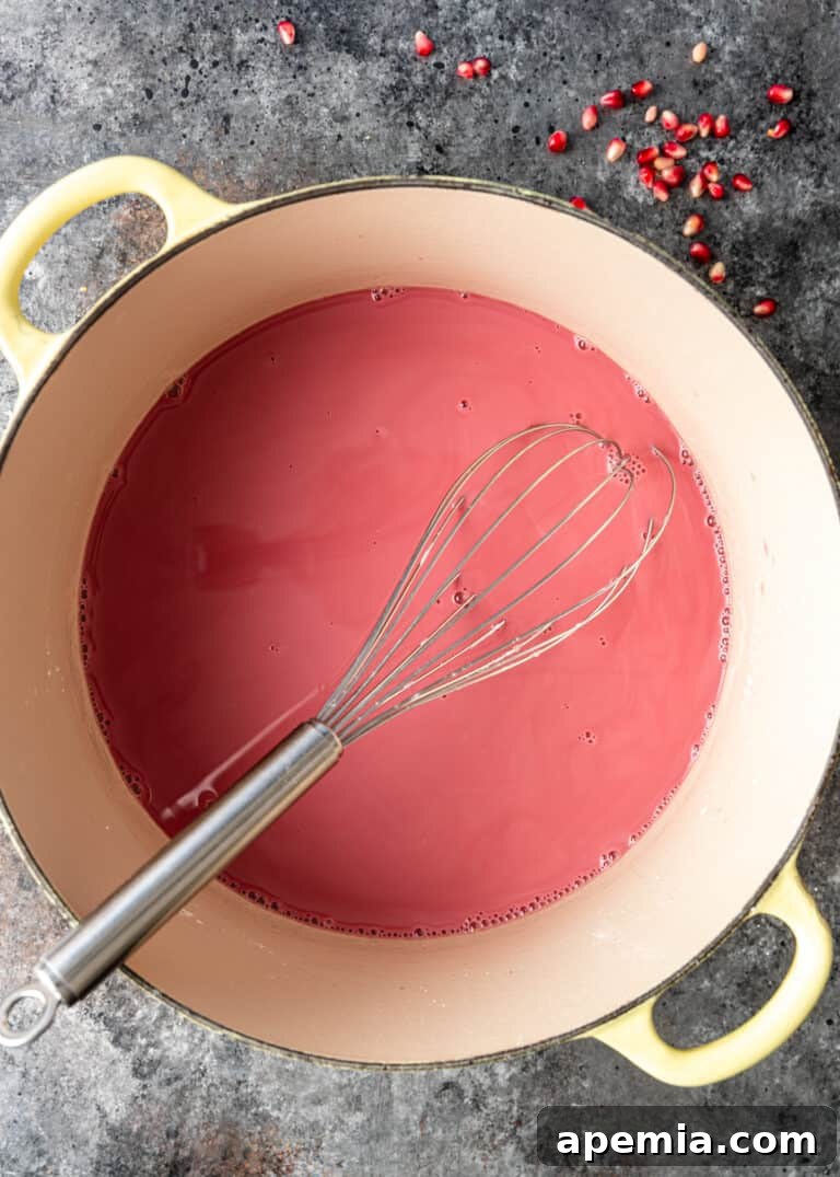 The vibrant pink pomegranate mixture thickening in a Dutch oven, with a whisk demonstrating its consistency.