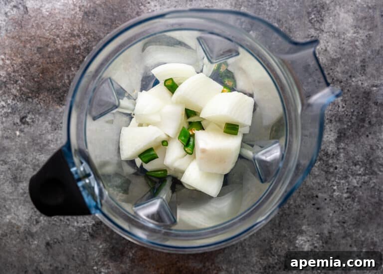 Overhead view of chopped white onion and green serrano chilies inside a blender, with a small amount of water.