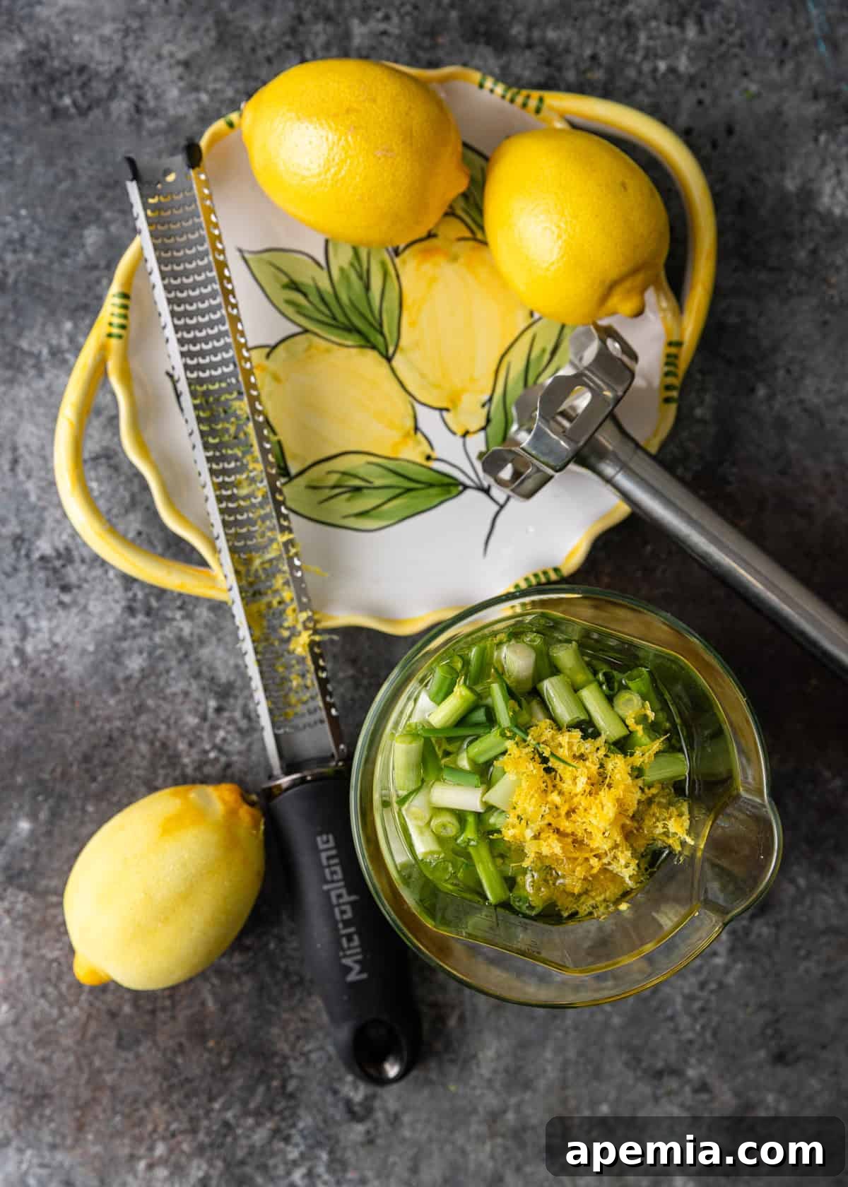 Overhead process shot demonstrating lemon zesting: a microplane zester rests on a platter with two whole lemons, while a third lemon, already zested, lies next to a blender containing initial dressing ingredients.