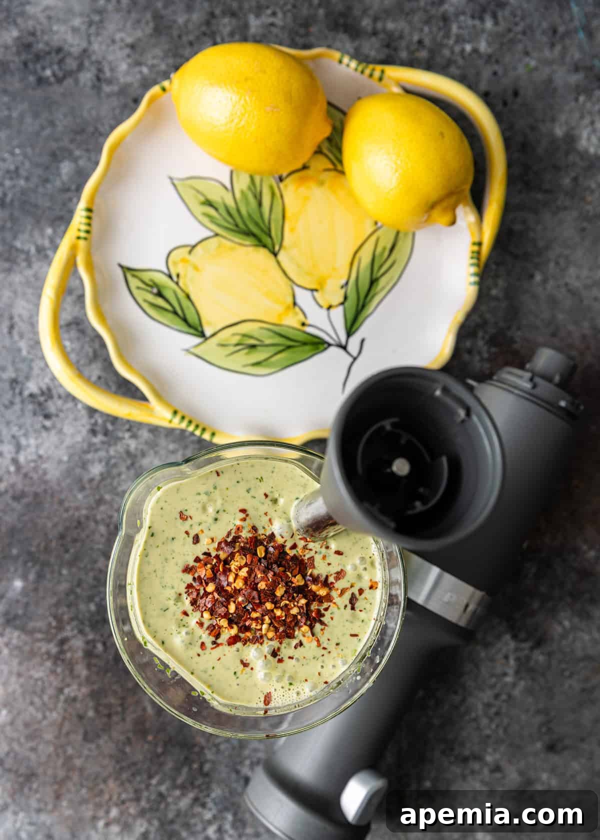 Overhead process shot of lemon tahini dressing ingredients mixed in a blender, with two fresh lemons on a platter to the side, indicating the dressing is nearly complete.
