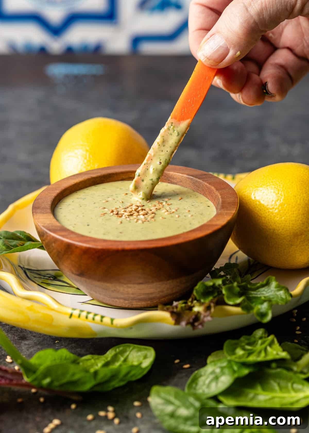 Closeup side view of a hand dipping a roasted carrot into a wooden bowl filled with creamy lemon tahini salad dressing, set on a platter with two fresh lemons.
