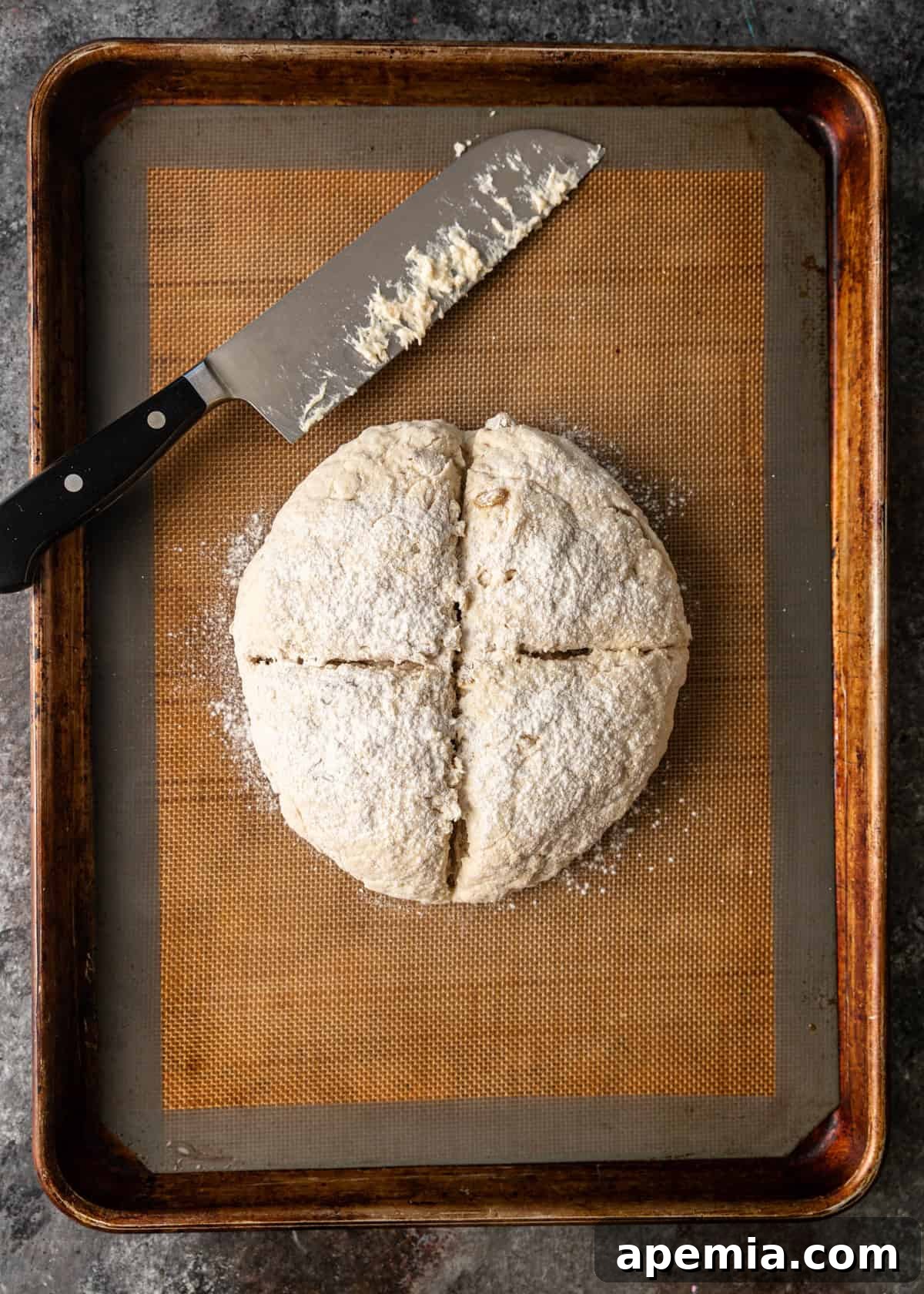 Overhead process shot of the flour-dusted, scored soda bread dough, perfectly shaped into a ball and placed on a baking sheet, ready for the oven.