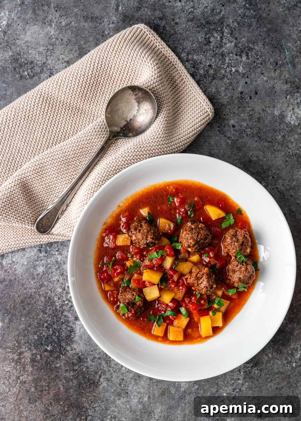Overhead view of a pristine white bowl of Ras Asfour meatball stew, perfectly garnished, with a cloth napkin and spoon neatly arranged beside it.