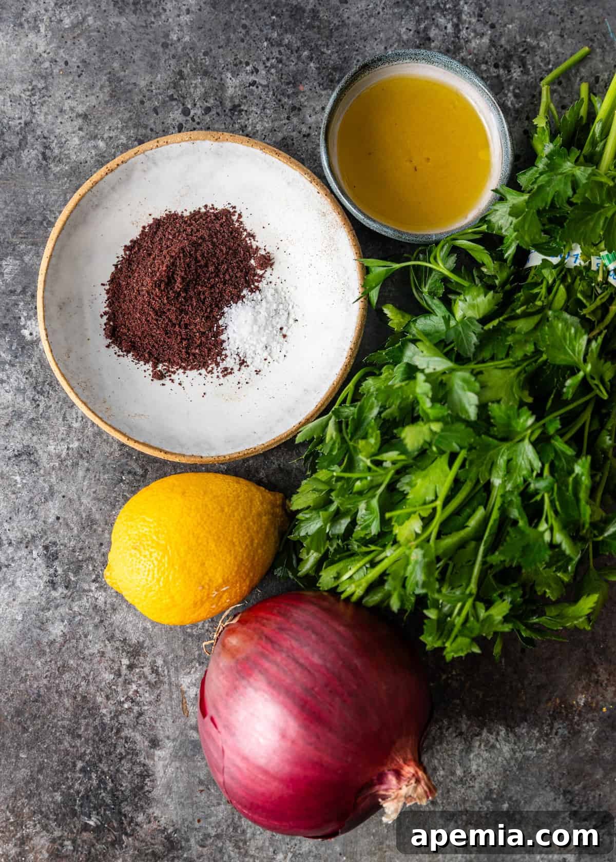 Overhead shot of fresh ingredients laid out on a cutting board, including a red onion, lemon, sumac, and parsley.