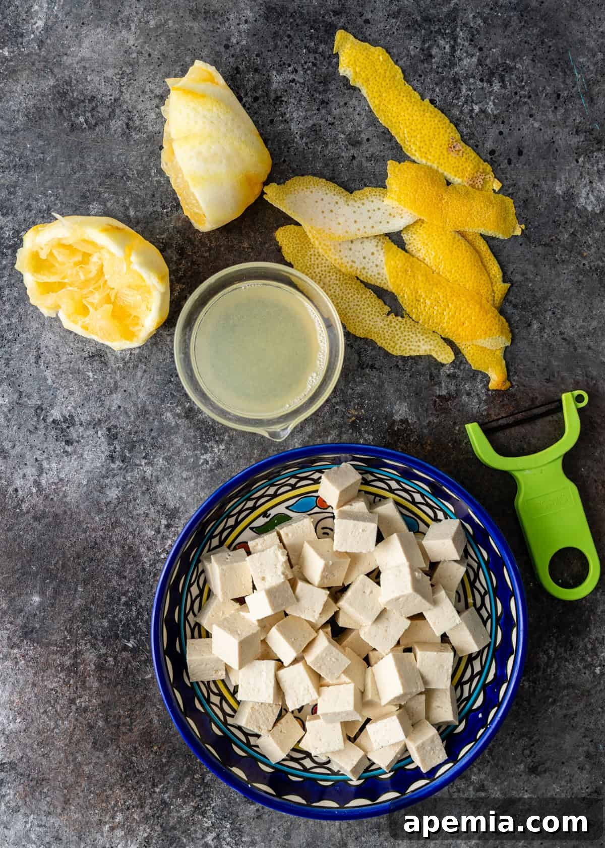 overhead process shot: bowl of chopped tofu and lemon juice in measuring cup with peeler and lemon skins on side