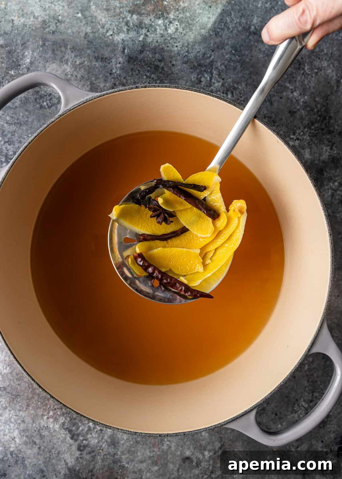 overhead process shot: large stock pot with chicken stock, soy sauce, star anise, and spoon holding red pepper flakes and the peels of the lemon over top