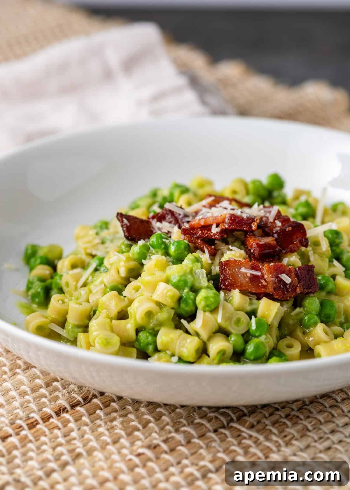 closeup side view: pasta e piselli on plate topped with bacon and parmesan cheese