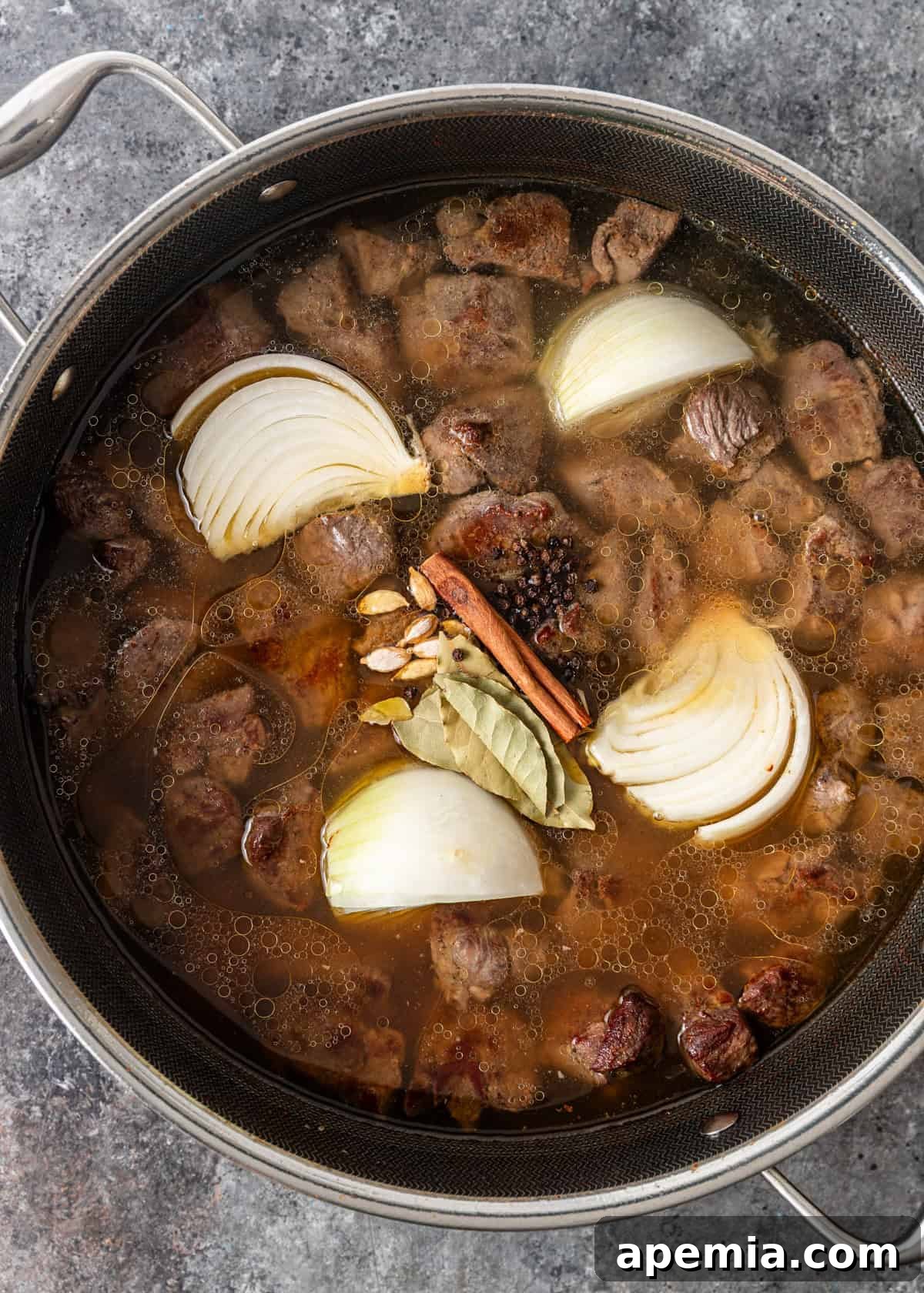 overhead process shot: water, cardamom, bay leaves, cinnamon, peppercorns, and onion added to cooked lamb