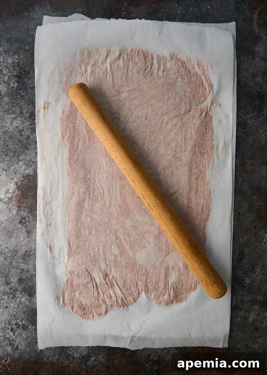 overhead process shot: meat rolled between two parchment papers with rolling pin on top