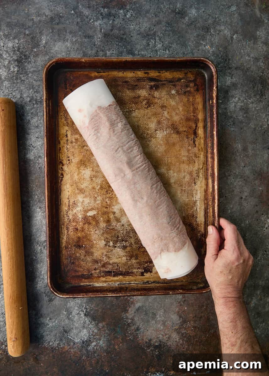 overhead process shot: hand holding sheet pan with rolled meat in parchment paper on top with rolling pin on the side